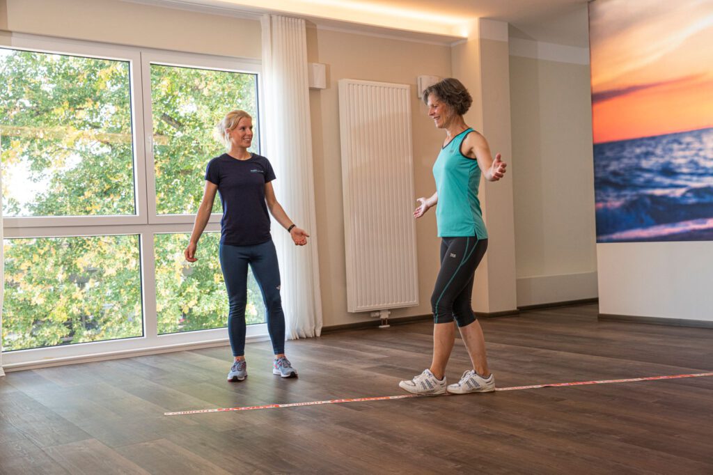 Ältere Frau balanciert bei einer Gleichgewichtsübung auf einer Bodenlinie, Trainerin Elli Schulte leitet an